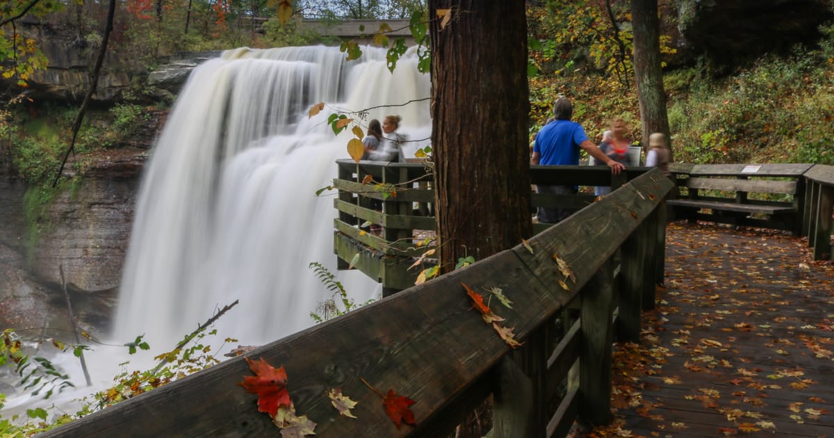 A powerful waterfall cascades over layered rock ledges into a misty gorge, viewed from a wooden observation deck surrounded by autumn foliage in warm oranges, reds, and greens. The long-exposure silky water contrasts beautifully with the rustic…