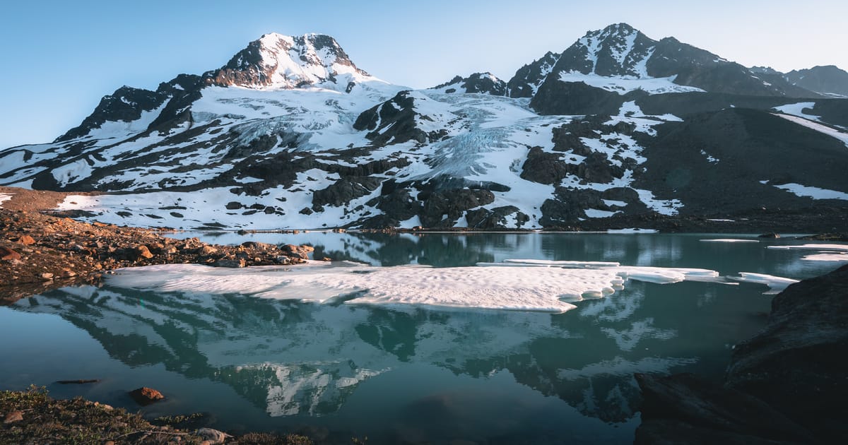 A glacially-fed alpine lake reflects snow-capped mountain peaks and a glacier in still, turquoise waters, with floating ice floes in the foreground and warm early-morning or late-evening light kissing the rocky shoreline.