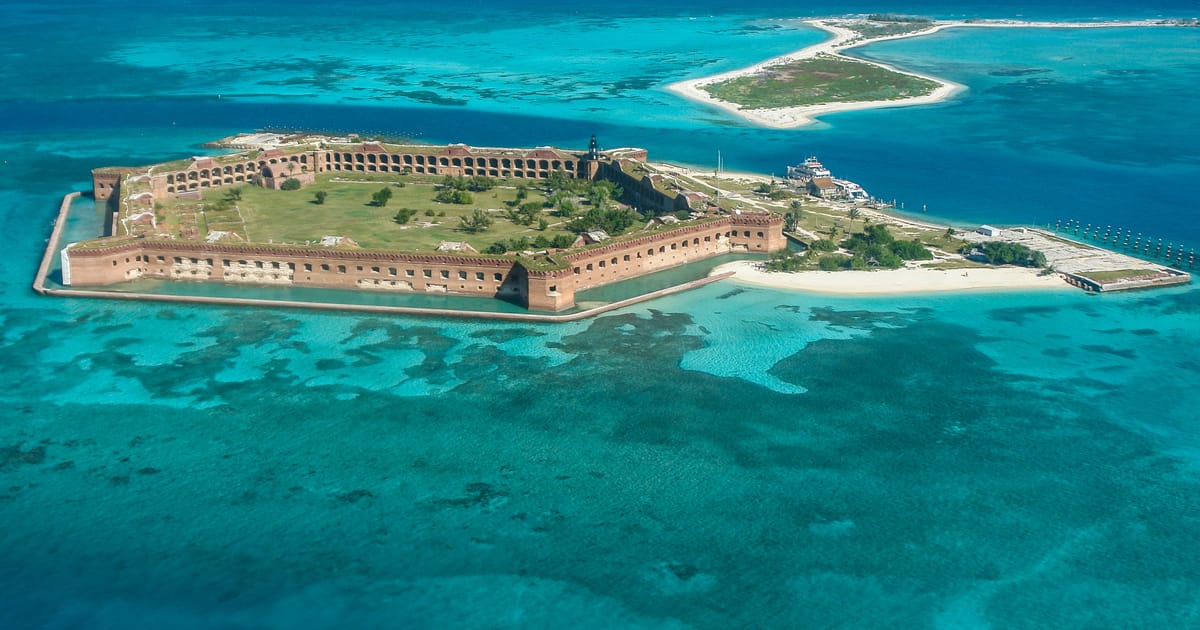 An aerial view of Fort Jefferson on Garden Key in Dry Tortugas National Park, Florida, surrounded by vivid turquoise and teal shallow waters with visible coral reefs. The massive hexagonal 19th-century brick fort sits on a small island under bright…