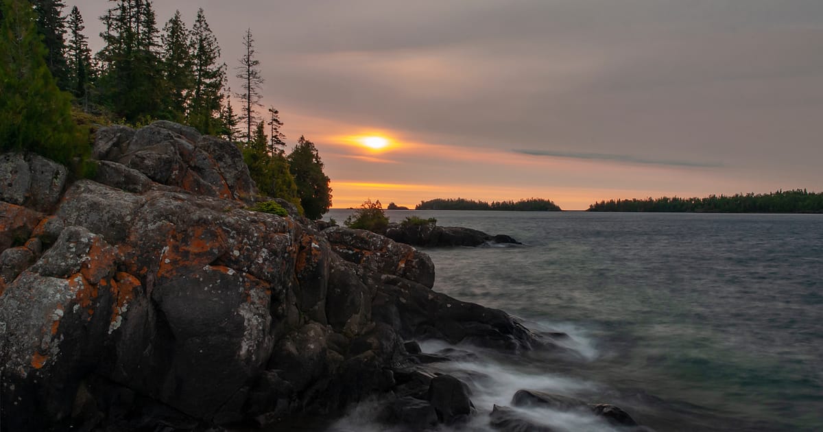 A dramatic sunset over a large lake viewed from a rocky, lichen-covered shoreline lined with boreal conifers, with forested islands silhouetted in the distance. The long-exposure capture renders the lapping water silky and ethereal beneath a hazy,…