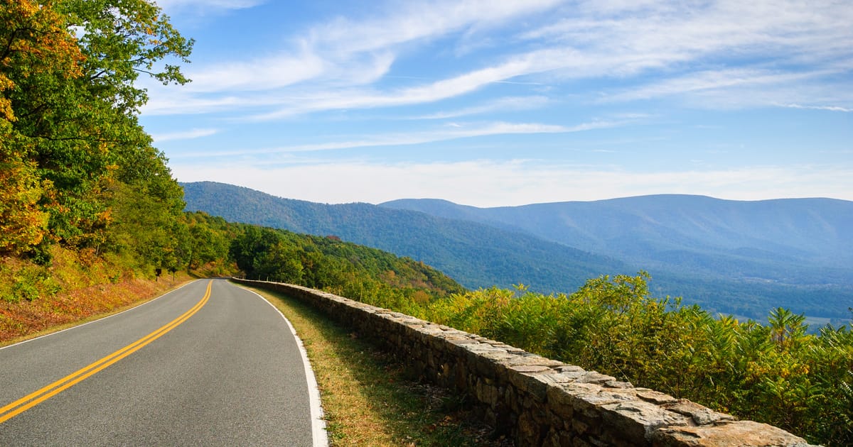 A scenic two-lane mountain road with a rustic stone guardrail curves along a ridgeline in Shenandoah National Park, with sweeping views of the Blue Ridge Mountains layered in soft haze under a bright autumn sky.