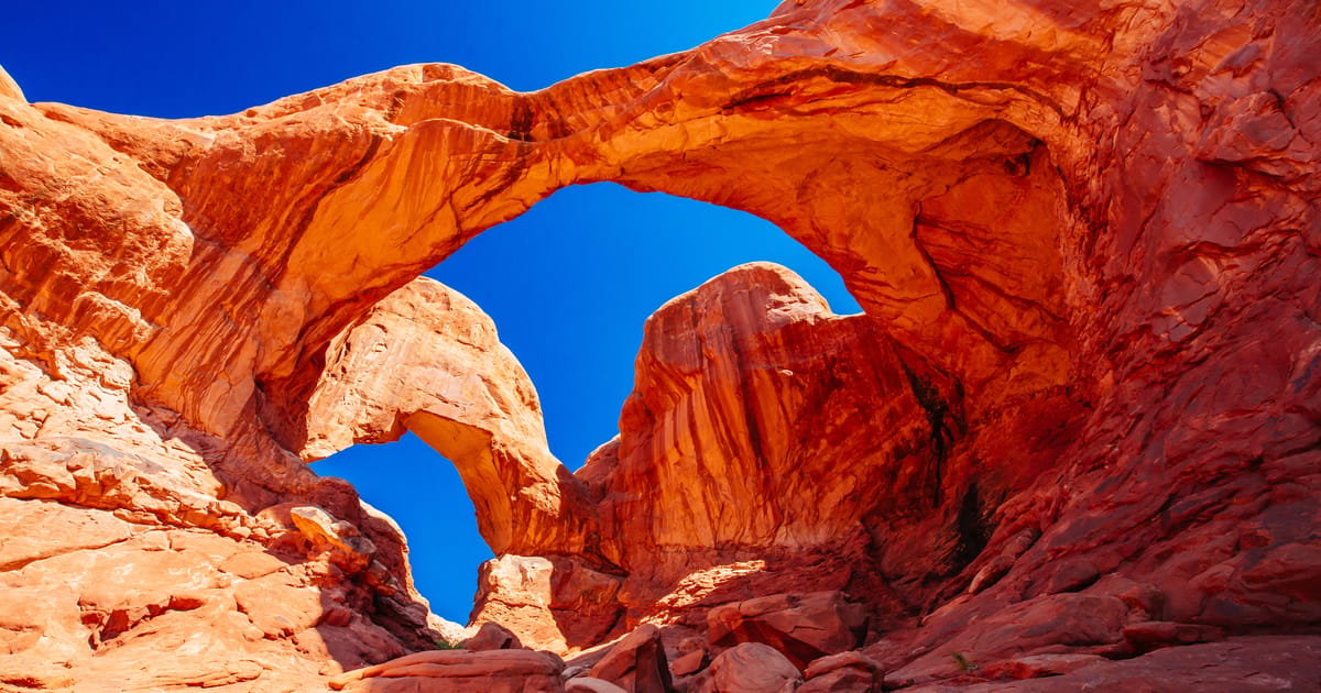 Double Arch at Arches National Park in Utah, two natural sandstone arches framing a vivid cobalt blue sky, photographed from a dramatic low-angle perspective that emphasizes the towering scale and warm orange-red hues of the Entrada sandstone.
