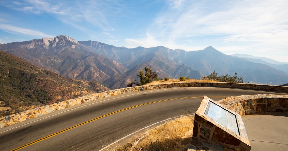 A winding mountain road curves along a stone-walled scenic overlook with a wayside exhibit panel, offering a sweeping view of rugged Sierra Nevada peaks and canyon ridgelines under a bright blue sky with wispy clouds.