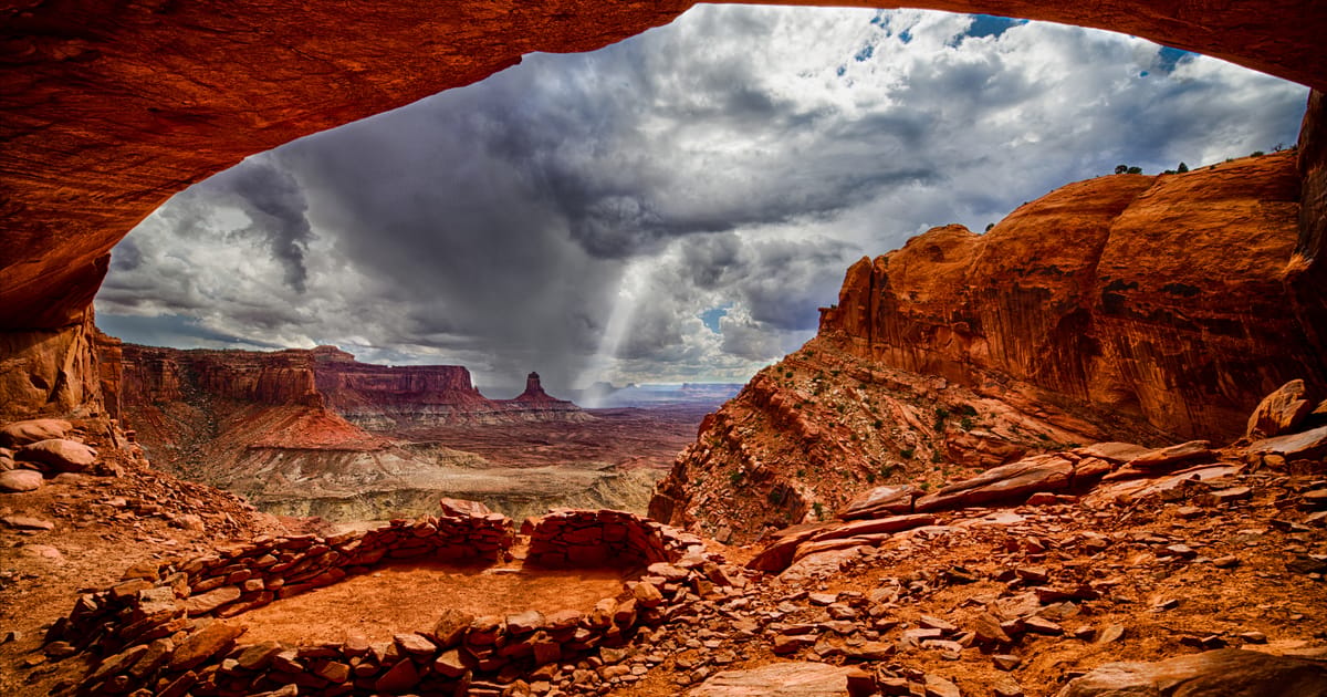 A dramatic view from inside a natural sandstone alcove at False Kiva in Canyonlands National Park, revealing ancient stone ruins in the foreground and sweeping canyon mesas under a stormy, cloud-laden sky. The warm red-orange tones of the rock…