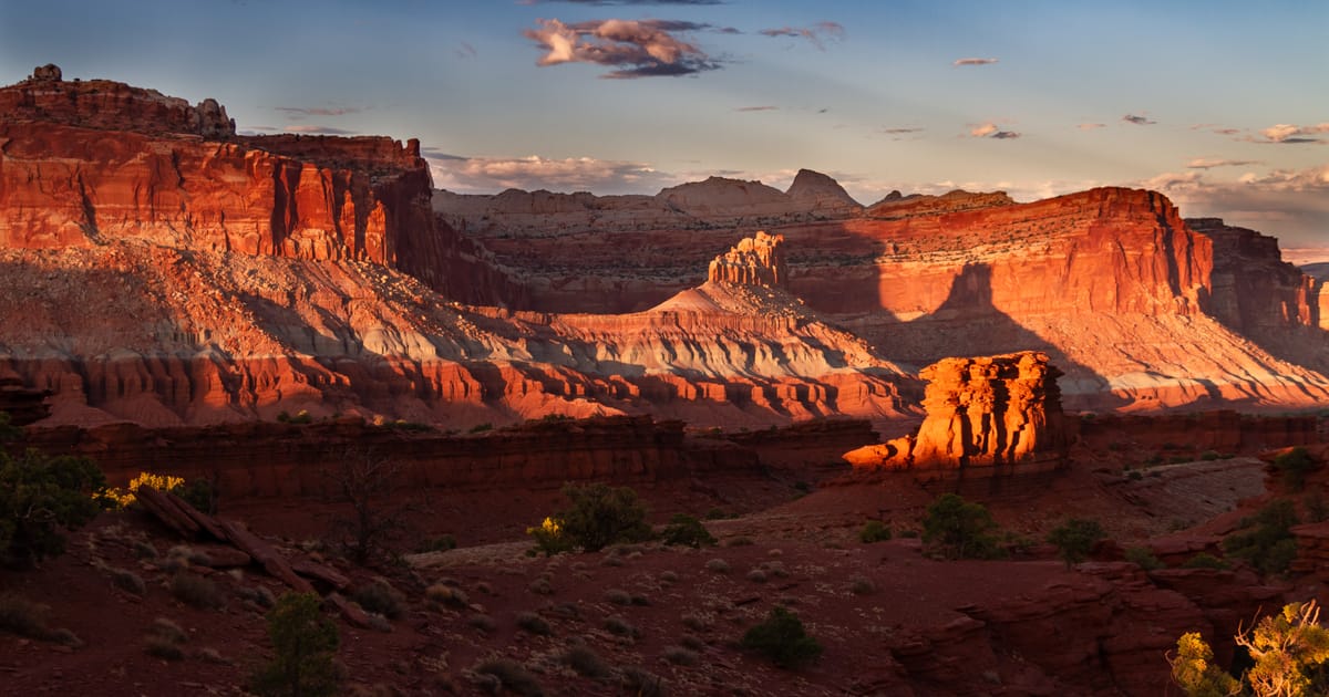 A sweeping panorama of red sandstone mesas and layered canyon walls bathed in warm golden-hour light, likely Capitol Reef National Park in Utah, with dramatic shadows and glowing orange cliffs set against a blue sky with scattered clouds.