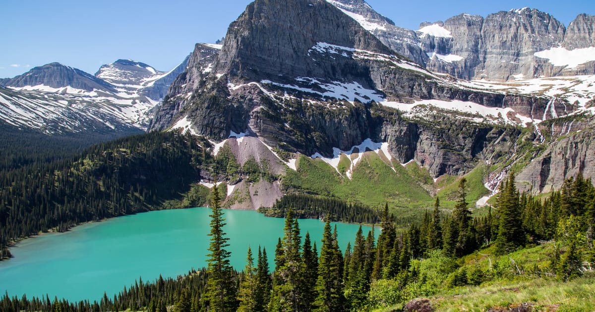 A stunning alpine lake with vivid turquoise glacial water nestled among dense evergreen forests and dramatic snow-dusted rocky peaks, photographed under bright midday sunlight with crisp blue skies conveying a sense of grandeur and wilderness.