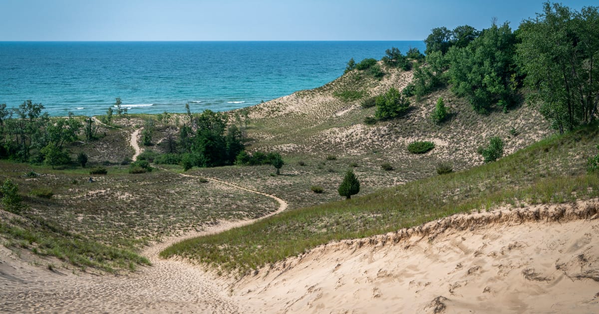 A sweeping view of sandy dunes with sparse vegetation and a winding trail leading down toward the vivid turquoise-blue waters of Lake Michigan, bathed in bright midday summer light with a clear sky.