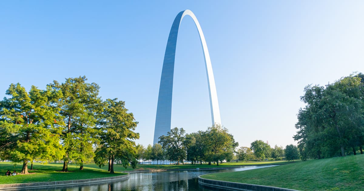 The iconic Gateway Arch in St. Louis rises dramatically against a clear blue sky, reflected in a calm reflecting pool surrounded by lush green parkland. Warm morning light illuminates the stainless steel structure and verdant trees, creating a…