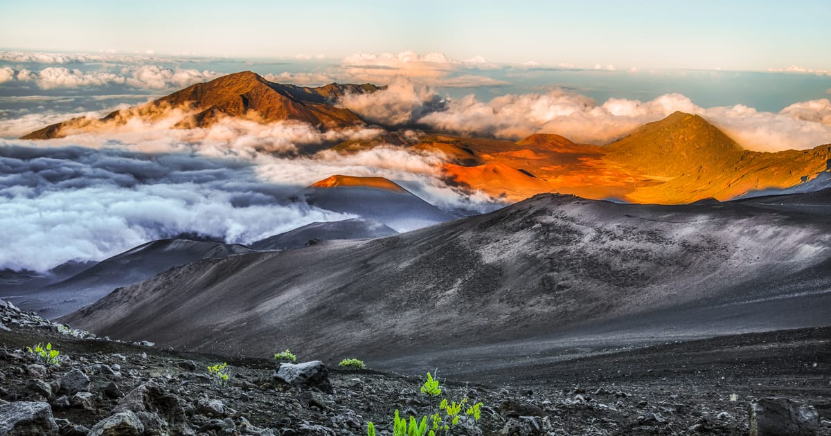 A dramatic aerial view of Haleakalā volcanic crater on Maui, Hawaii, with cinder cones bathed in warm golden-orange sunrise light surrounded by a sea of clouds. The stark dark volcanic terrain in the foreground contrasts with vibrant illuminated…