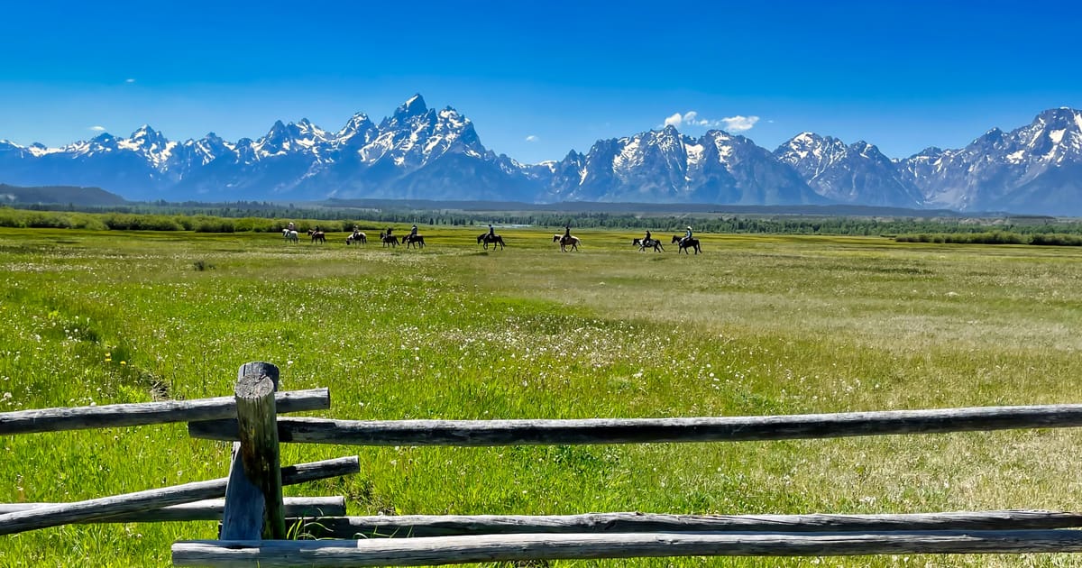 A sweeping summer meadow in Grand Teton National Park features a group of horseback riders crossing a vast green valley, with the iconic jagged, snow-capped Teton Range dramatically filling the horizon under a brilliant blue sky. A weathered…