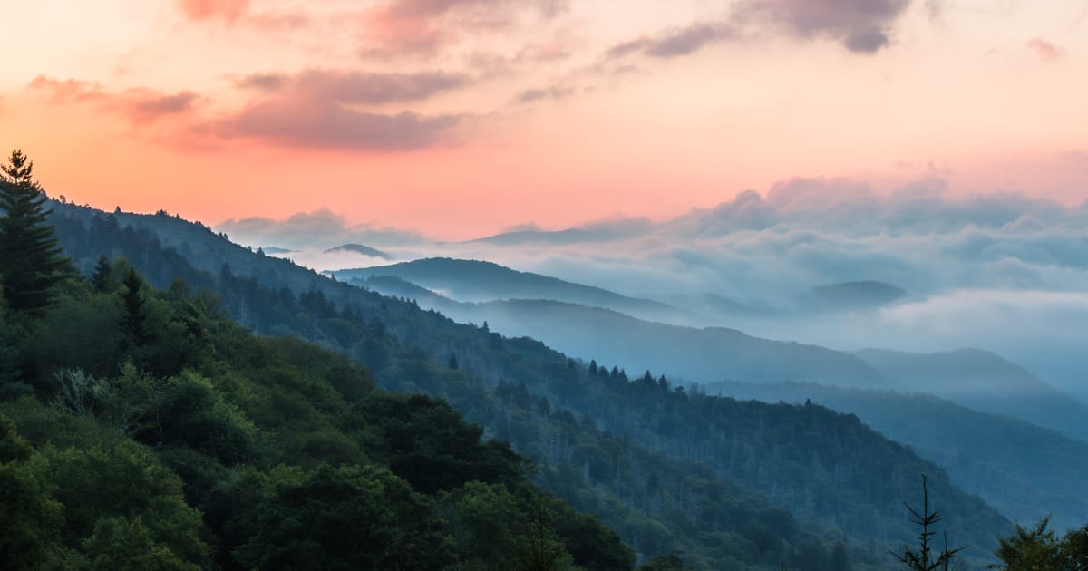 A sweeping panoramic view of layered, mist-filled mountain valleys covered in dense deciduous and evergreen forest, captured at sunrise or sunset with a dramatic pink and orange sky punctuated by soft clouds and flowing fog banks between the…
