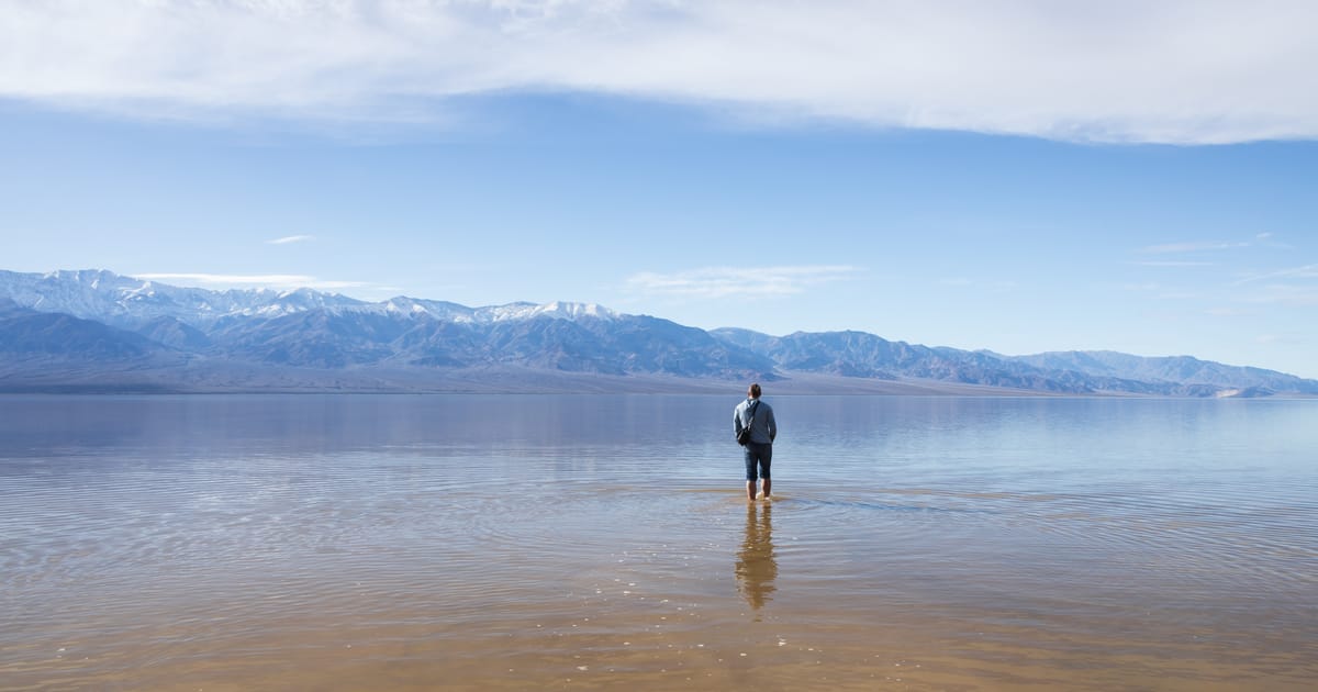 A lone figure wades through the shallow, mirror-like waters of a flooded desert basin — likely Badwater Basin in Death Valley National Park — with snow-dusted mountain ranges stretching across the horizon under a expansive blue sky with wispy…