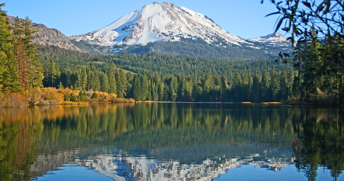 A snow-capped volcanic peak — likely Lassen Peak in Lassen Volcanic National Park, California — rises above a dense evergreen forest, with its reflection mirrored perfectly in the calm, glassy surface of Manzanita Lake. The autumn foliage adds warm…