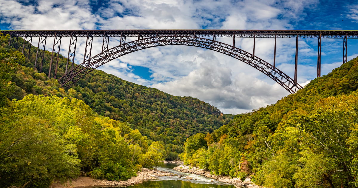The iconic New River Gorge Bridge, a massive steel arch bridge spanning the densely forested gorge above the New River in West Virginia, photographed from below with lush green canopy and partly cloudy blue skies creating a dramatic, majestic mood.