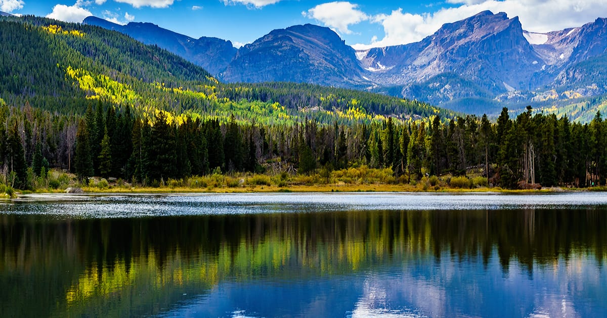 A serene alpine lake in Rocky Mountain National Park reflects the surrounding evergreen forest and rugged mountain peaks under a partly cloudy blue sky. The scene captures early autumn foliage with golden aspen trees dotting the hillsides, creating…