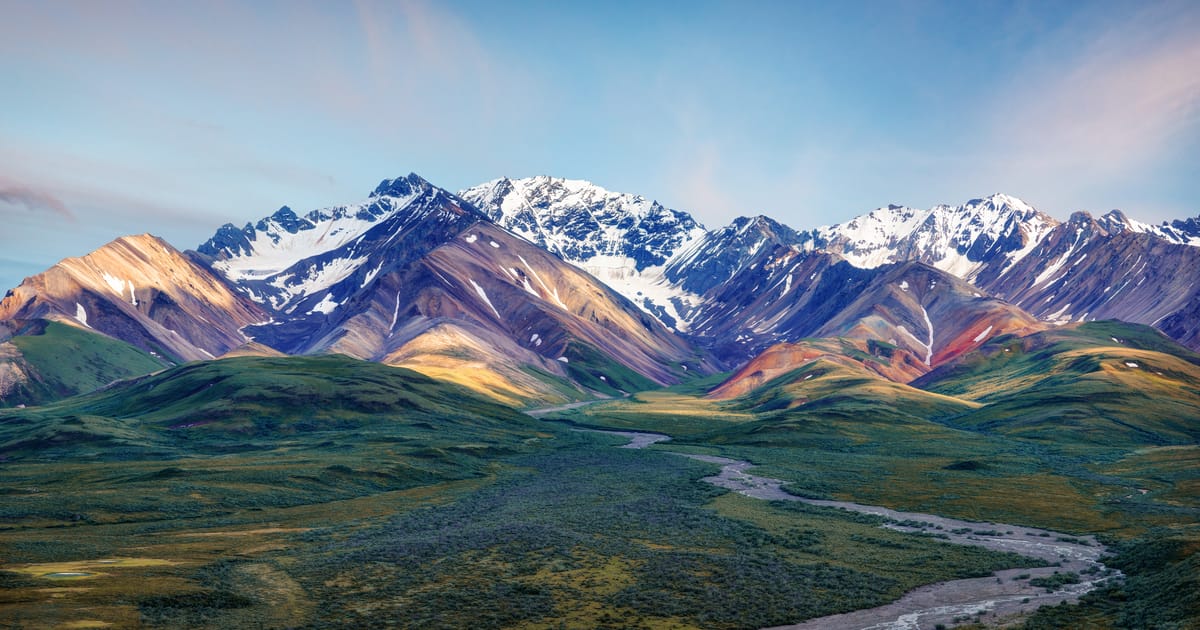 A sweeping panoramic view of the Alaska Range in Denali National Park, featuring snow-capped peaks with colorful mineral-streaked slopes in warm amber, rust, and olive tones, above a braided glacial river winding through lush green tundra valleys.…