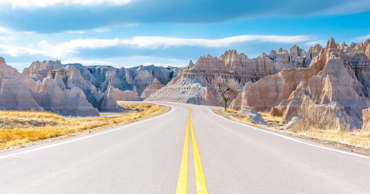 A two-lane highway with double yellow center lines winds through the dramatic eroded buttes and spires of Badlands National Park, South Dakota, under a partly cloudy blue sky. The scene has an open, inviting mood with warm golden grasses flanking…