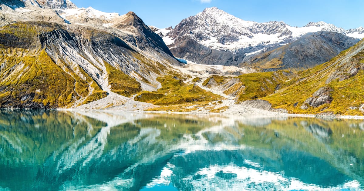 A stunning alpine lake with vivid turquoise water perfectly mirrors snow-capped mountain peaks and glaciers in calm, glassy conditions. Bright midday light illuminates golden-green tundra slopes, rocky moraines, and dramatic glaciated summits under…