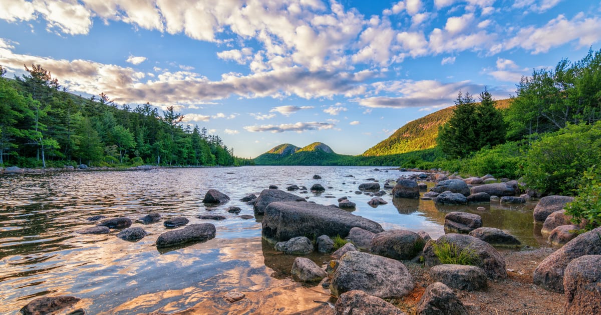 Jordan Pond in Acadia National Park, Maine, featuring smooth glacial boulders along the shoreline with the iconic Bubbles mountains rising in the background, bathed in warm golden-hour light against a dramatic blue sky filled with altocumulus clouds.