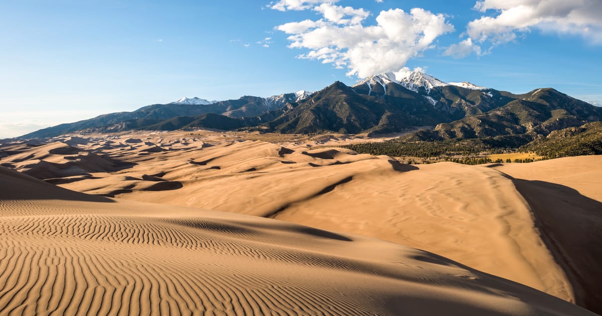 Great Sand Dunes National Park in Colorado showcases towering rippled sand dunes in the foreground with snow-capped Sangre de Cristo Mountains rising dramatically in the background. Warm golden sunlight illuminates the dune ridges, creating striking…