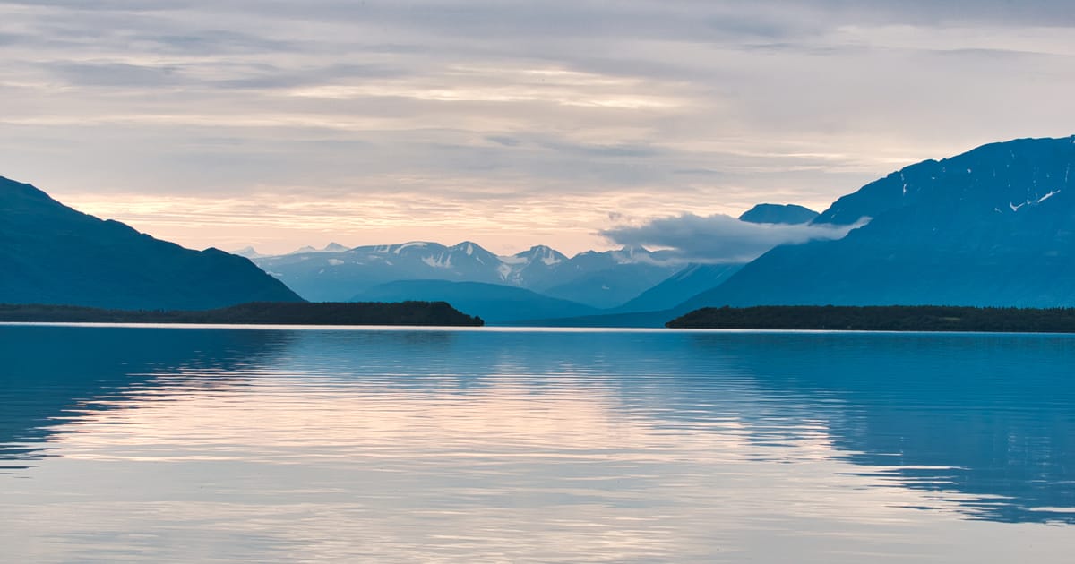 A serene glacial lake reflects soft pastel twilight hues beneath a layered overcast sky, framed by dark silhouetted mountains and snow-capped peaks receding into the misty distance. The calm, mirror-like water and cool blue tones create a tranquil,…
