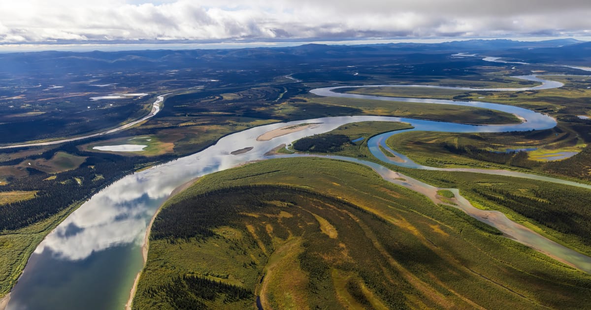 An aerial view of a vast braided and meandering river system winding through boreal forest and tundra flats, likely the Yukon or Kobuk River in Alaska, under dramatic partly cloudy skies with soft diffused light reflecting off the calm water.