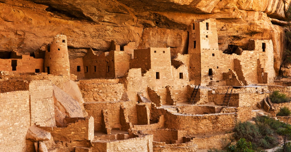 Cliff Palace at Mesa Verde National Park, an ancient Ancestral Puebloan cliff dwelling nestled beneath a massive sandstone overhang, bathed in warm golden afternoon light that accentuates the ochre and tan tones of the multi-story stone structures.