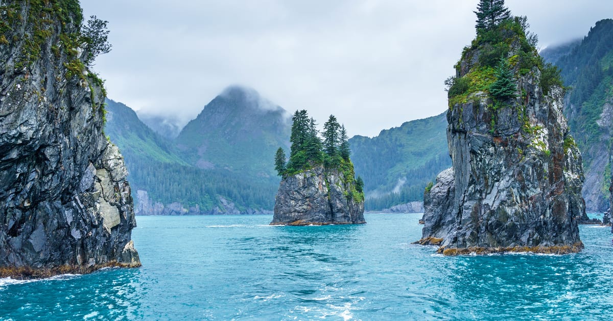Dramatic sea stacks and rocky spires rise from vivid turquoise waters in what appears to be Kenai Fjords National Park, Alaska, with evergreen trees clinging to the summits and misty, cloud-shrouded mountains receding into the background. The…