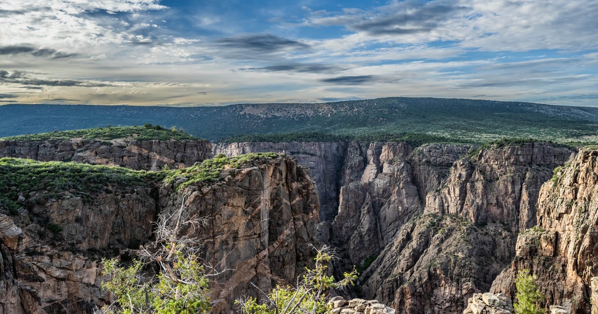 A dramatic aerial view of the Black Canyon of the Gunnison, featuring sheer dark metamorphic rock walls plunging into a deep narrow gorge, under a partially cloudy blue sky with warm afternoon light illuminating the canyon rim.