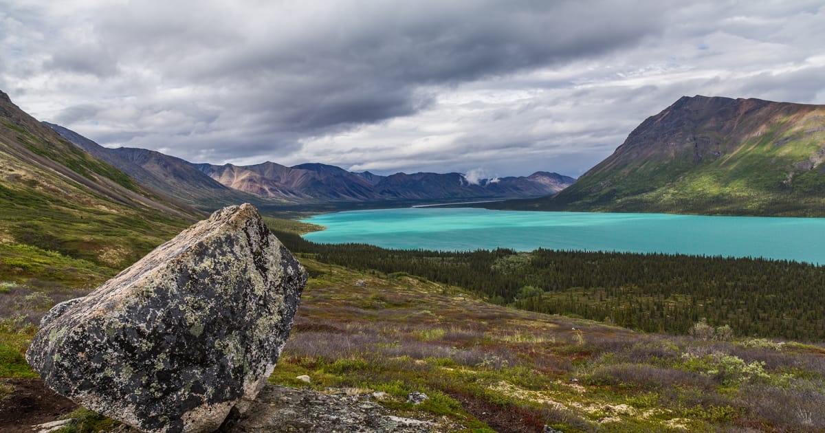 A striking glacial lake with vivid turquoise water nestled between rugged mountain ranges, framed by a large lichen-covered boulder in the foreground and a dense boreal forest below under dramatic overcast skies.
