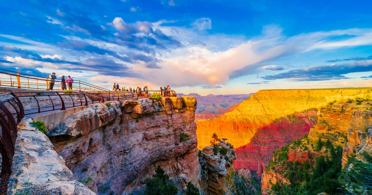 A dramatic sunset view of the Grand Canyon from Mather Point overlook on the South Rim, with warm golden and red light bathing the layered sandstone cliffs while tourists gather along the railed viewing platform.