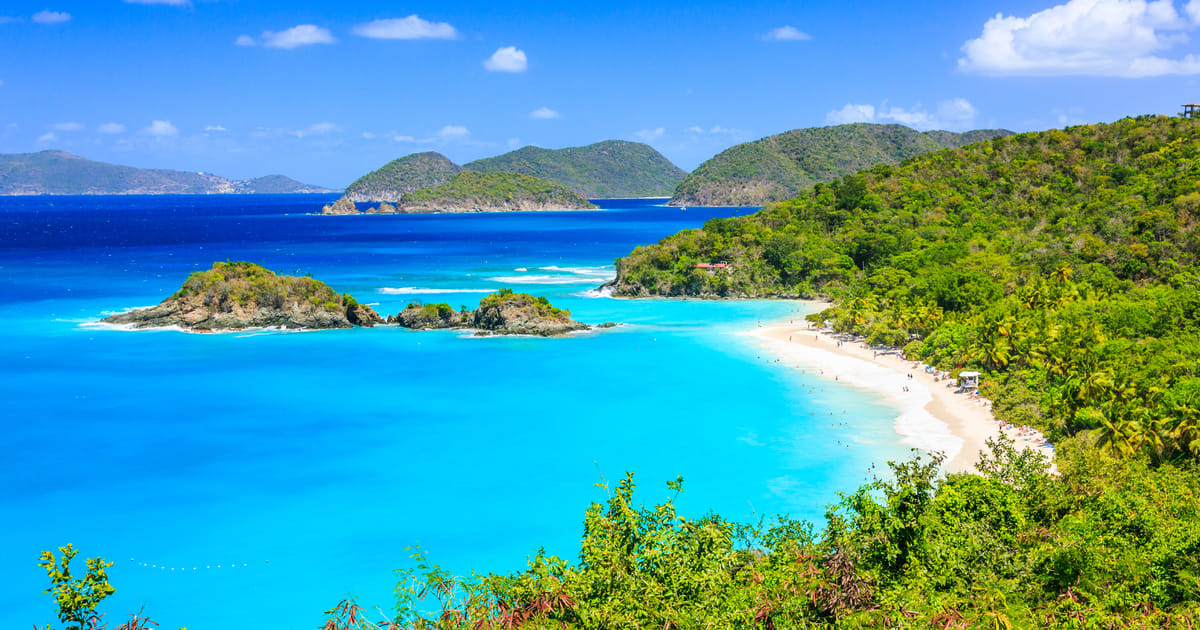 A stunning aerial view of Trunk Bay in St. John, US Virgin Islands National Park, featuring vivid turquoise and deep blue waters curving around a white sandy beach flanked by lush tropical hillsides. The scene is bathed in brilliant midday sunlight,…