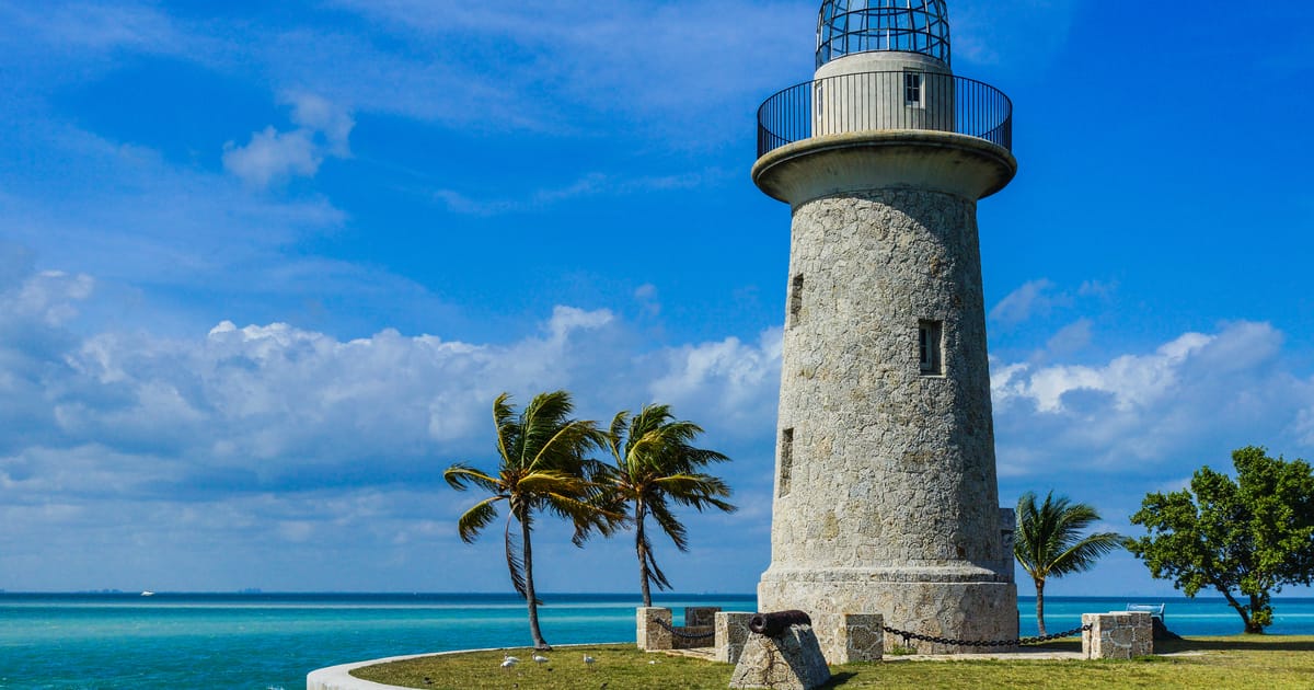 The Boca Chita Key lighthouse in Biscayne National Park stands on a small coral island surrounded by turquoise waters, with palm trees swaying in the breeze under a vibrant blue sky with scattered clouds. The historic stone tower is bathed in bright…