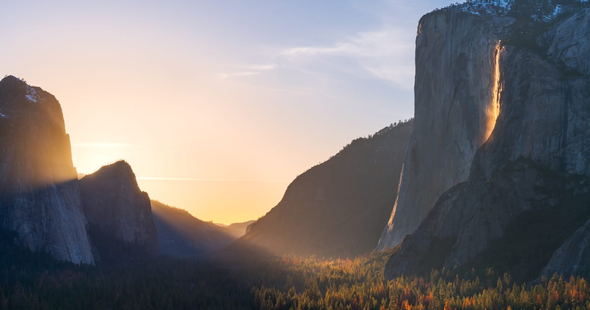 Yosemite Valley at golden hour featuring El Capitan's sheer granite face with Horsetail Fall glowing orange in the famous 'Firefall' effect, framed by forested valley floor with autumn foliage and a warm sunset illuminating misty air between canyon…