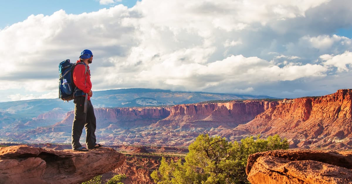 A hiker with a blue backpack and red jacket stands on a red sandstone ledge overlooking a sweeping canyon landscape at Capitol Reef National Park, with cloud-filled skies and warm afternoon light illuminating the layered cliffs.