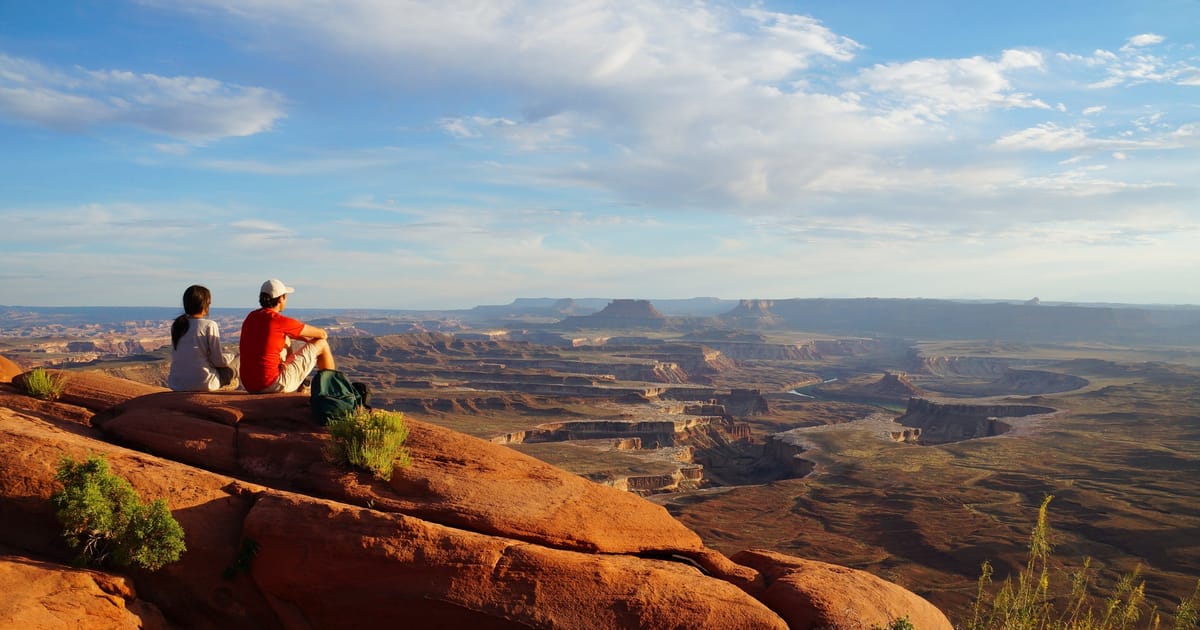 A hiker in a wide-brimmed hat traverses a narrow sandstone ledge trail in Canyonlands National Park, with sheer canyon walls dropping away on both sides and a vast red-rock mesa landscape extending to the horizon under a clear blue sky.