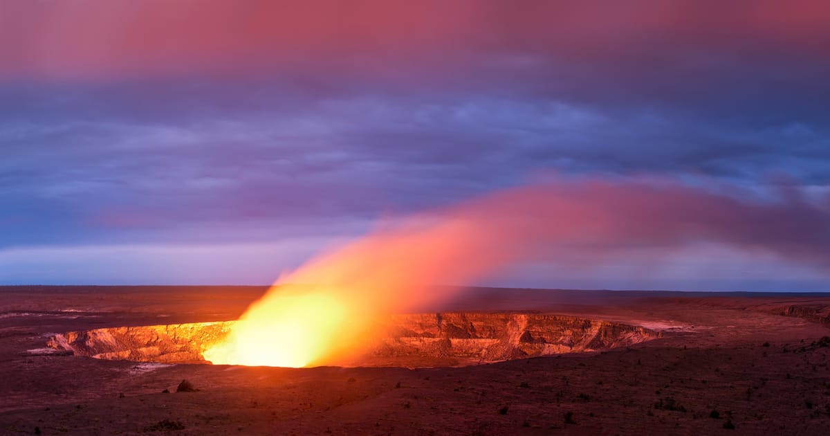 A glowing lava lake inside Halemaʻumaʻu Crater at Kīlauea volcano illuminates the caldera walls with intense orange and yellow light, casting a dramatic plume of volcanic gas across a twilight sky of deep blues and purples.