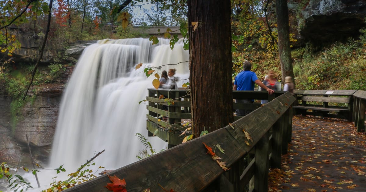 A powerful waterfall cascades over layered rock ledges into a misty gorge, viewed from a wooden observation deck surrounded by autumn foliage in warm oranges, reds, and greens. The long-exposure silky water contrasts beautifully with the rustic…