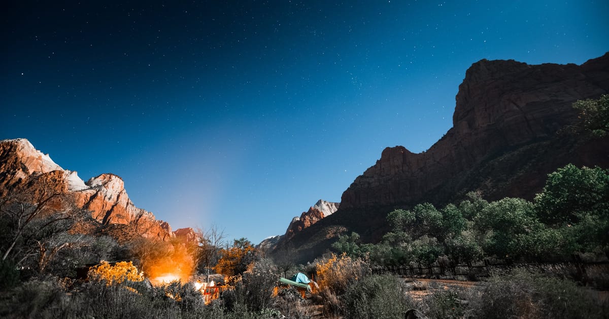 A canyon-floor campsite at dusk in Zion National Park, a campfire glowing beneath the sandstone walls.