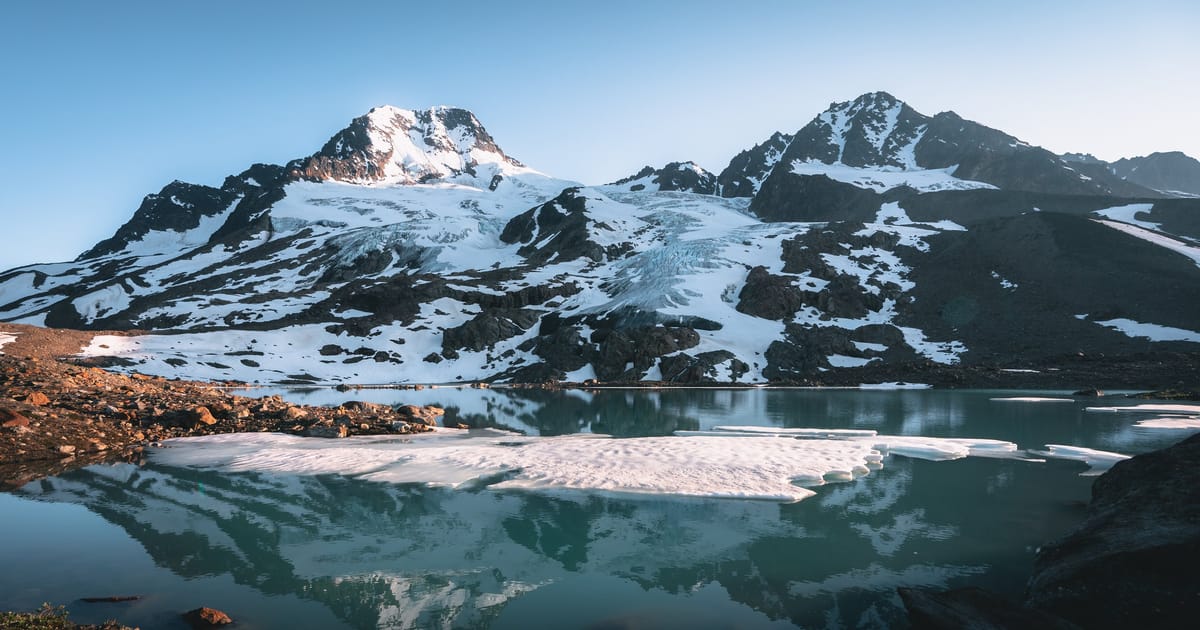 A glacially-fed alpine lake reflects snow-capped mountain peaks and a glacier in still, turquoise waters, with floating ice floes in the foreground and warm early-morning or late-evening light kissing the rocky shoreline.
