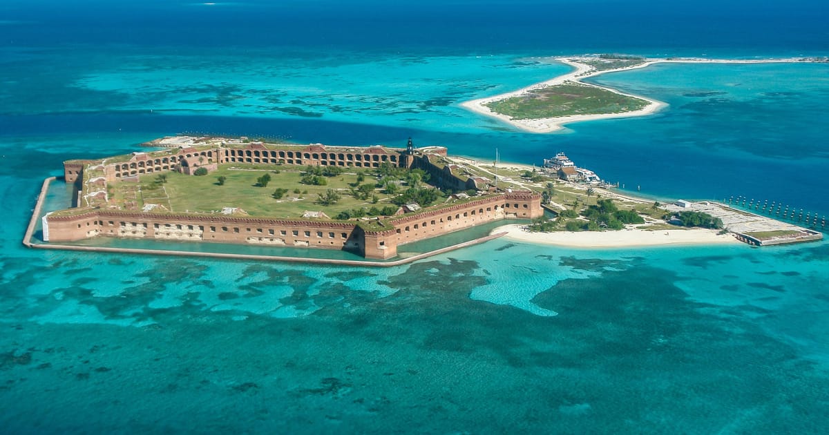An aerial view of Fort Jefferson on Garden Key in Dry Tortugas National Park, Florida, surrounded by vivid turquoise and teal shallow waters with visible coral reefs. The massive hexagonal 19th-century brick fort sits on a small island under bright…