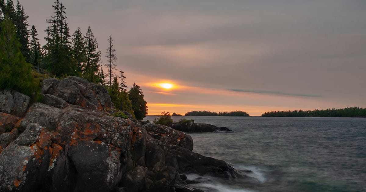 A dramatic sunset over a large lake viewed from a rocky, lichen-covered shoreline lined with boreal conifers, with forested islands silhouetted in the distance. The long-exposure capture renders the lapping water silky and ethereal beneath a hazy,…