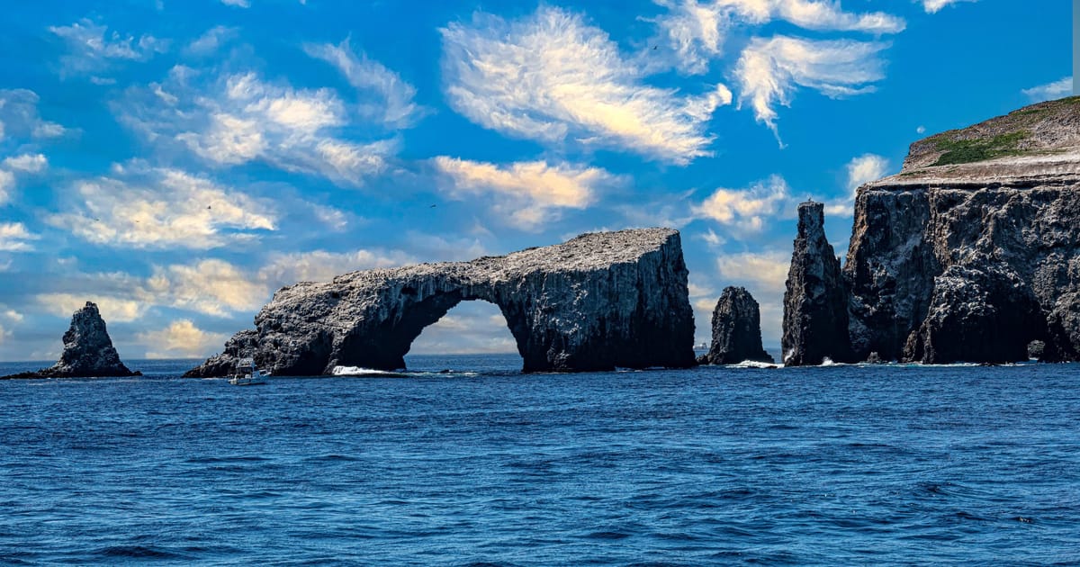 Anacapa Island's iconic Arch Rock in Channel Islands National Park, California — a weathered volcanic sea-arch rises from a deep blue Pacific alongside a slender rock spire, with sandstone island bluffs flanking the right and high cirrus clouds streaking a vivid summer sky.