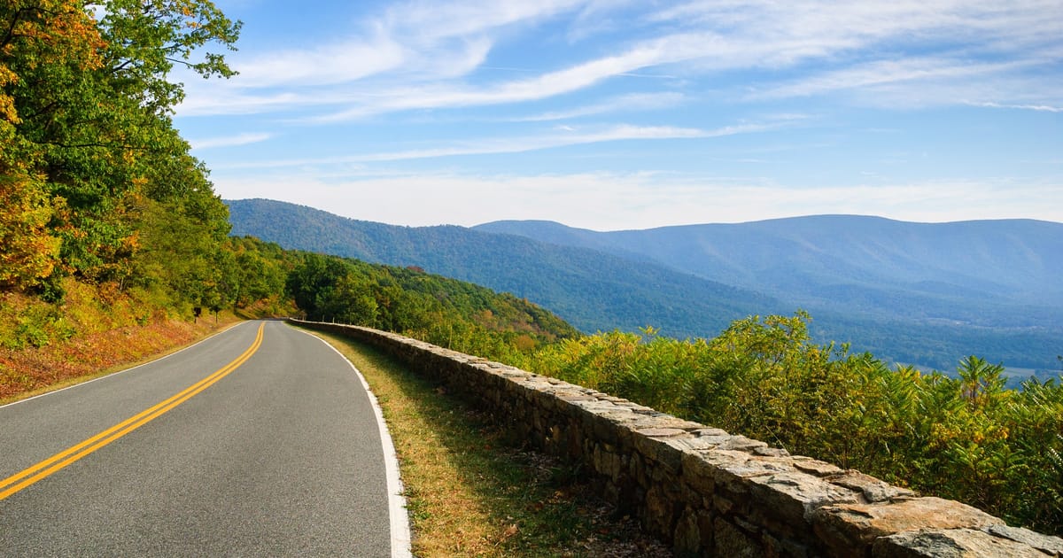 A scenic two-lane mountain road with a rustic stone guardrail curves along a ridgeline in Shenandoah National Park, with sweeping views of the Blue Ridge Mountains layered in soft haze under a bright autumn sky.