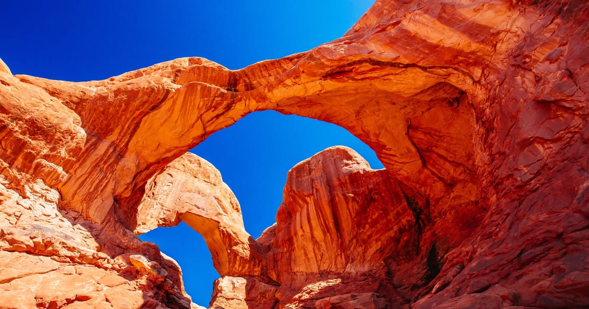 Double Arch at Arches National Park in Utah, two natural sandstone arches framing a vivid cobalt blue sky, photographed from a dramatic low-angle perspective that emphasizes the towering scale and warm orange-red hues of the Entrada sandstone.