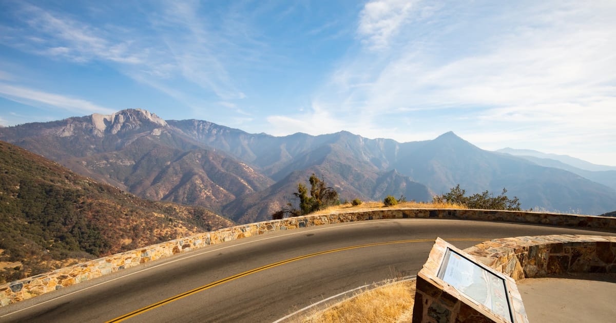 A winding mountain road curves along a stone-walled scenic overlook with a wayside exhibit panel, offering a sweeping view of rugged Sierra Nevada peaks and canyon ridgelines under a bright blue sky with wispy clouds.