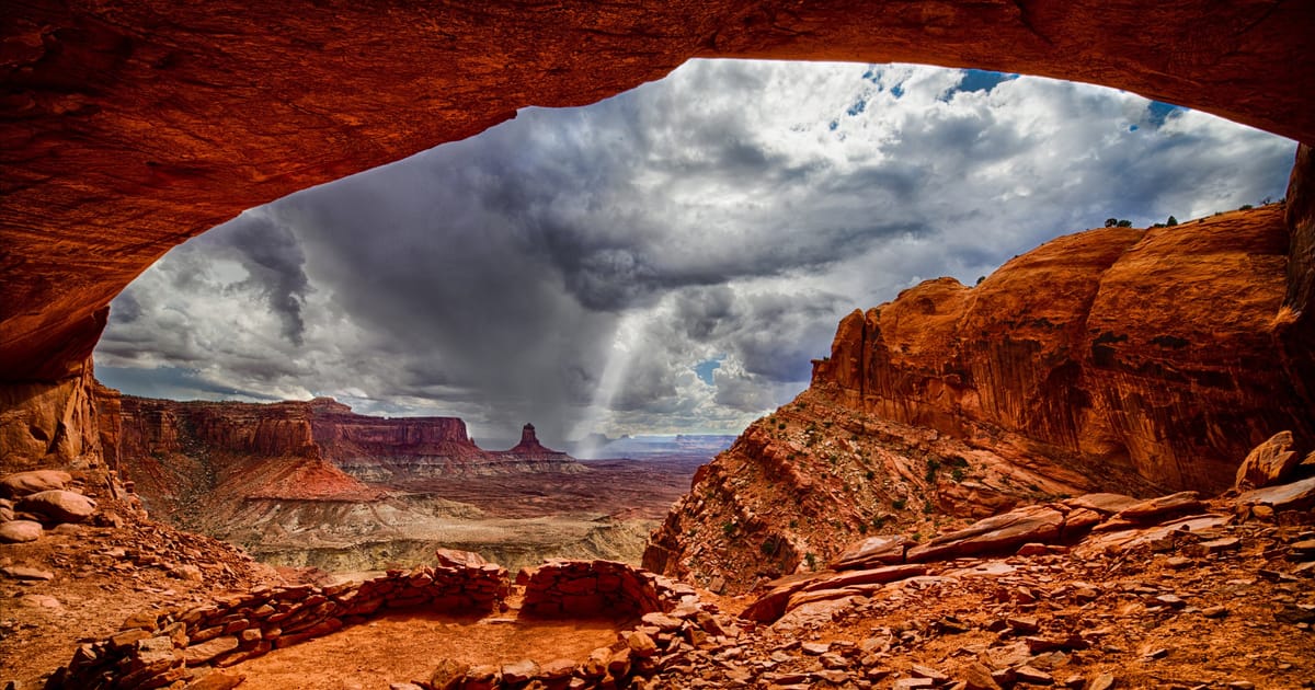 A dramatic view from inside a natural sandstone alcove at False Kiva in Canyonlands National Park, revealing ancient stone ruins in the foreground and sweeping canyon mesas under a stormy, cloud-laden sky. The warm red-orange tones of the rock…