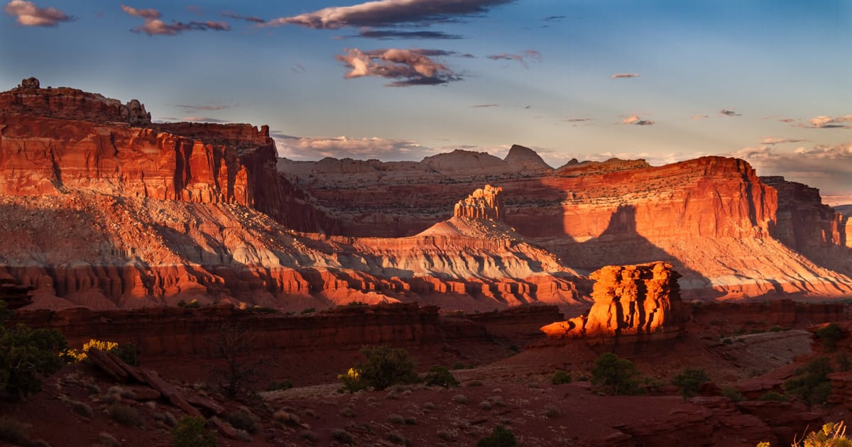 A sweeping panorama of red sandstone mesas and layered canyon walls bathed in warm golden-hour light, likely Capitol Reef National Park in Utah, with dramatic shadows and glowing orange cliffs set against a blue sky with scattered clouds.