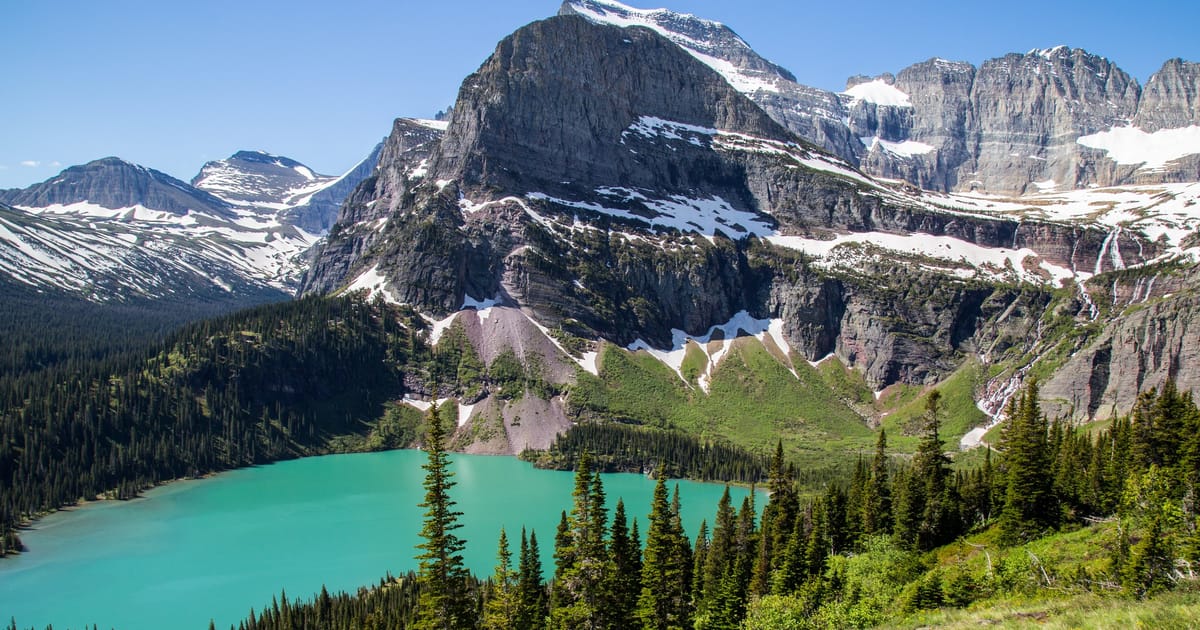 A stunning alpine lake with vivid turquoise glacial water nestled among dense evergreen forests and dramatic snow-dusted rocky peaks, photographed under bright midday sunlight with crisp blue skies conveying a sense of grandeur and wilderness.