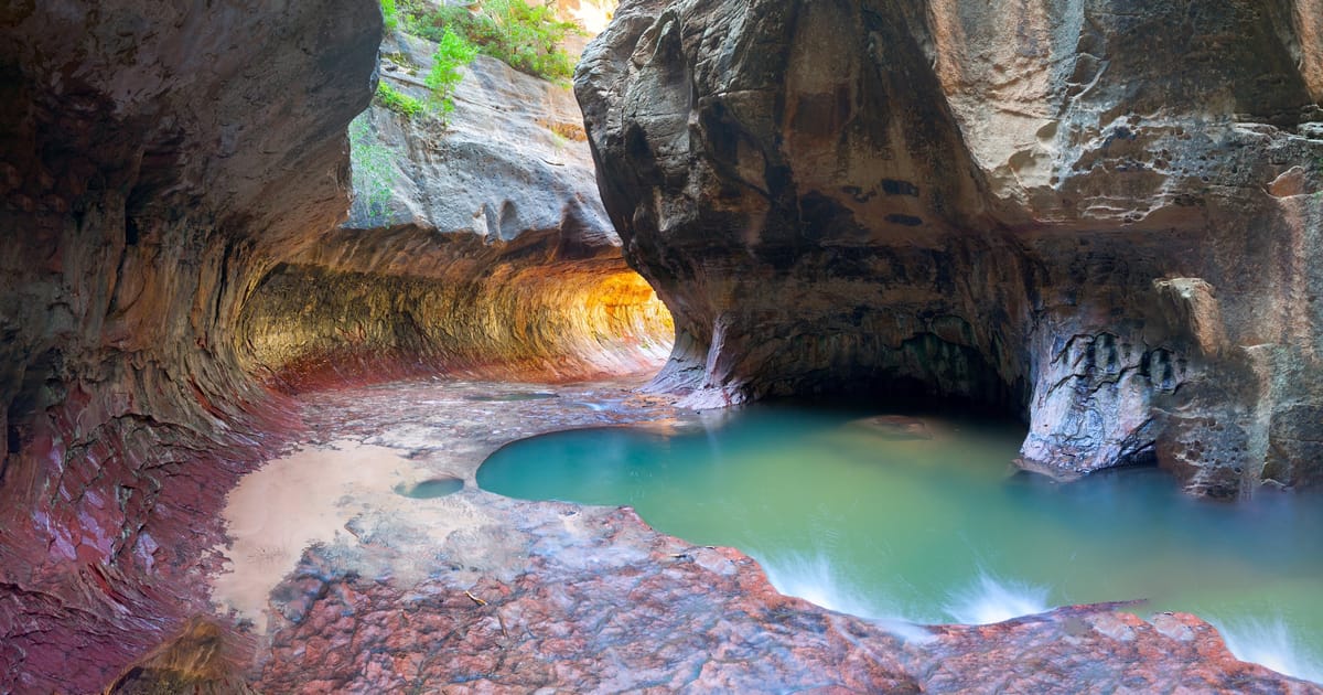 A stunning slot canyon interior known as 'The Subway' in Zion National Park, featuring smooth water-carved sandstone walls glowing with warm amber and rust tones, framing a serene turquoise pool below. The ethereal lighting filters through the…